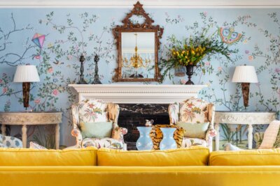 A bright living room in a Kappa Alpha Theta sorority house with floral wallpaper, a yellow sofa, two patterned armchairs, a fireplace, mirror, lamps, and vases with flowers on the mantel.