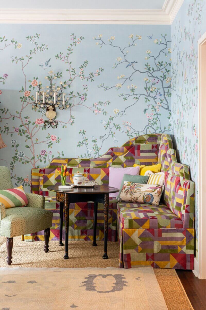 A colorful patterned banquette, small round black table with a tea set, green armchair, and floral wallpaper create a cozy, well-lit sitting area perfect for relaxing.