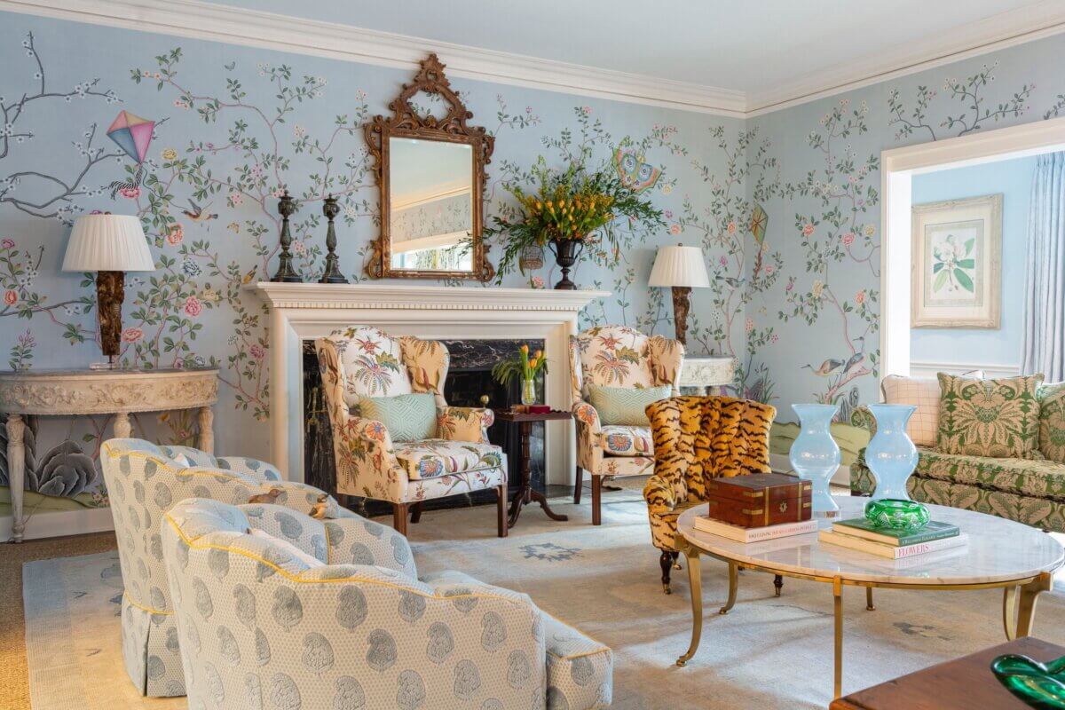 Elegant living room in this Kappa Alpha Theta sorority house features floral wallpaper, patterned armchairs, a decorative mirror above a white mantel, and a round coffee table with vases, books, and green glass decor.