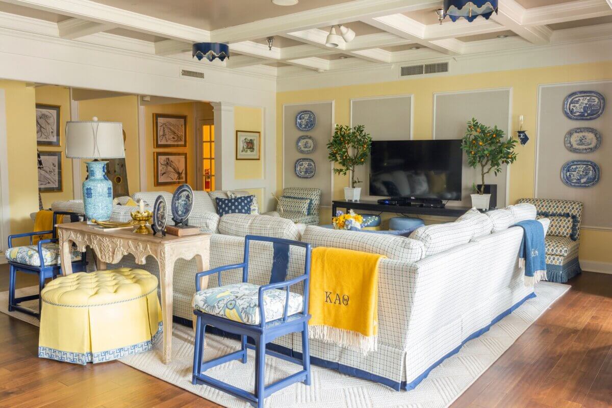 Bright living room in this Kappa Alpha Theta sorority house features patterned sofas, blue chairs, yellow accents, a large TV, decorative plates on the wall, plants, and framed artwork.