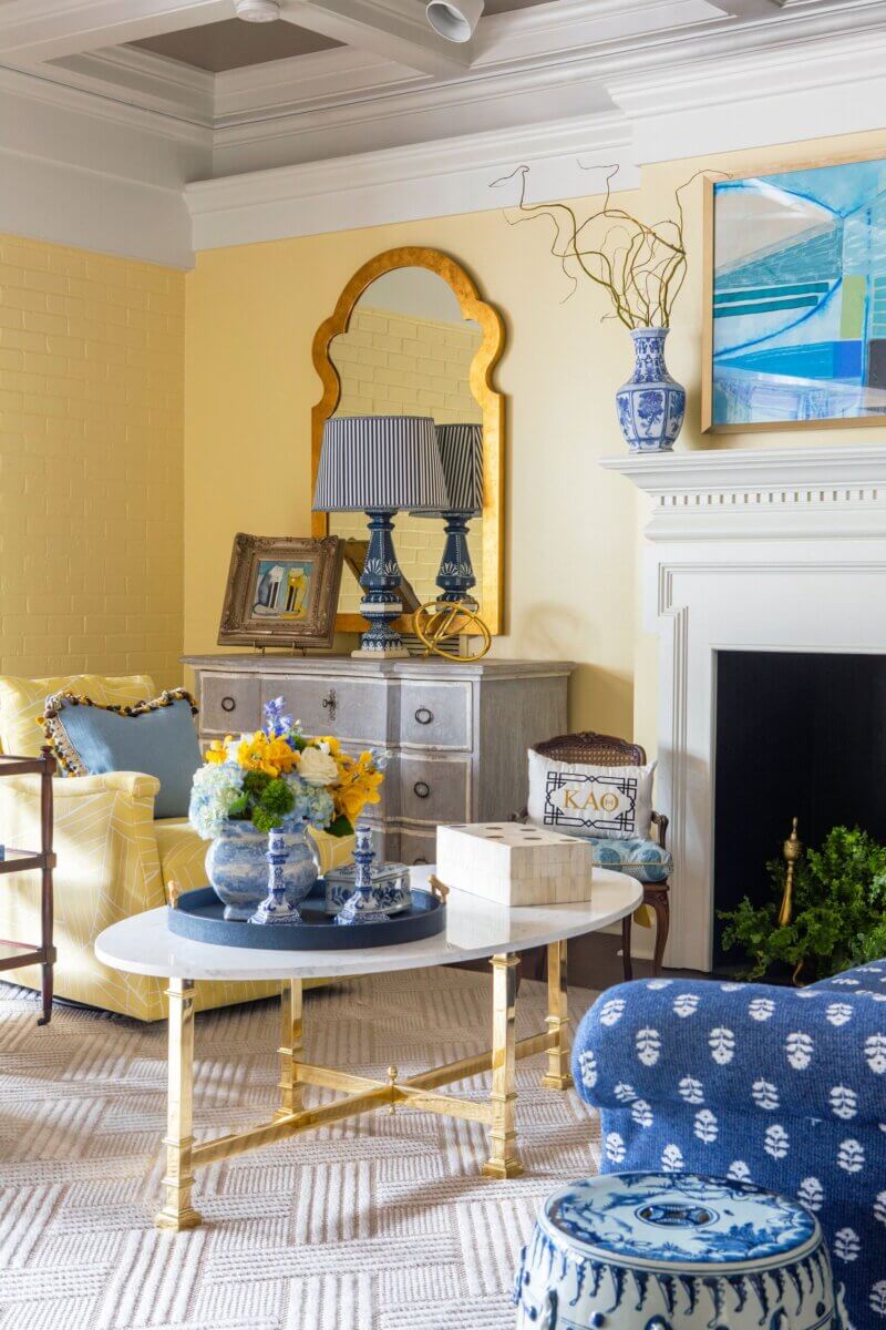A stylish living room in a sorority house features yellow walls, blue and yellow furniture, a gold-framed mirror, blue-and-white vases, and a round coffee table with decorative items reflecting Kappa Alpha Theta spirit.