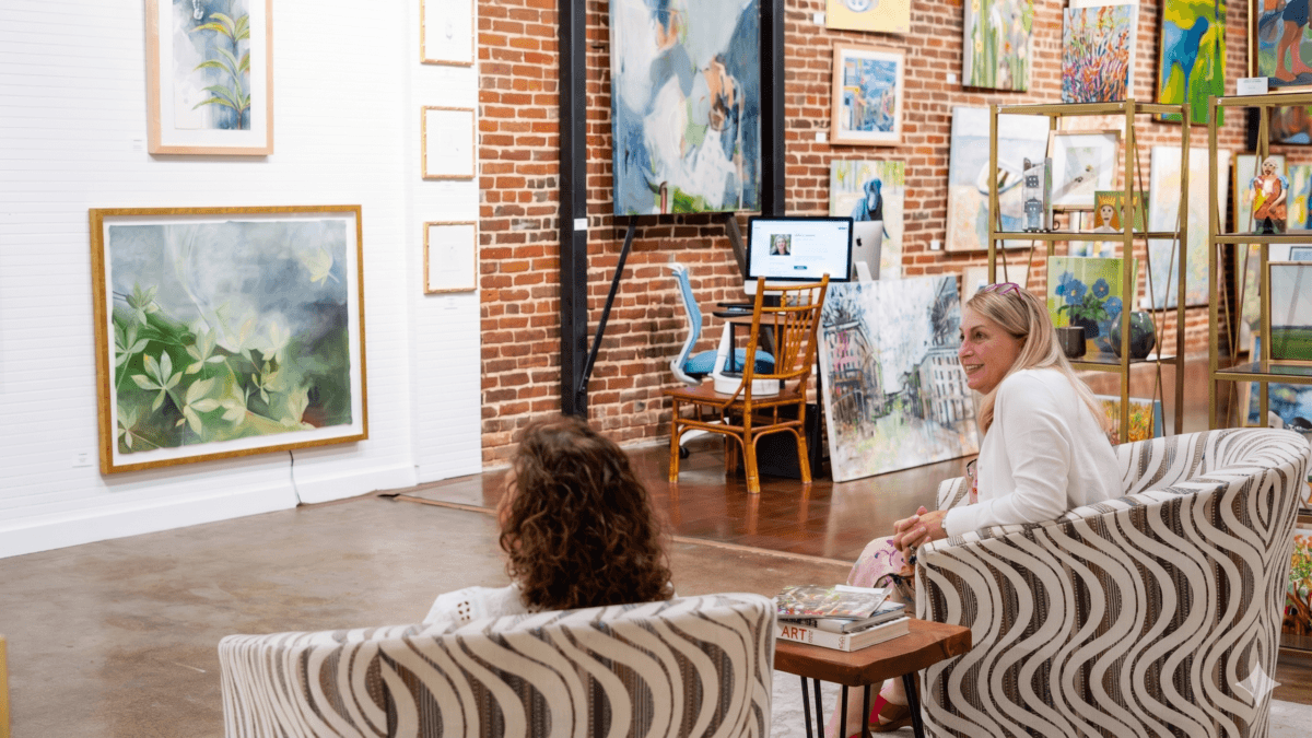 Two people sit in patterned chairs, chatting in an art gallery with brick walls and colorful paintings—a perfect spot if you're exploring things to do in Tupelo MS. A desk with a computer is visible in the background.