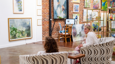 Two people sit in patterned chairs, chatting in an art gallery with brick walls and colorful paintings—a perfect spot if you're exploring things to do in Tupelo MS. A desk with a computer is visible in the background.