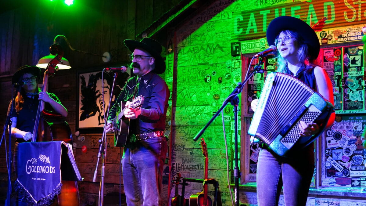 Three musicians perform on stage in Tupelo MS; one plays upright bass, another plays guitar and sings, and the third plays accordion. Posters and green lighting set the scene for a perfect summer getaway filled with music and travel vibes.