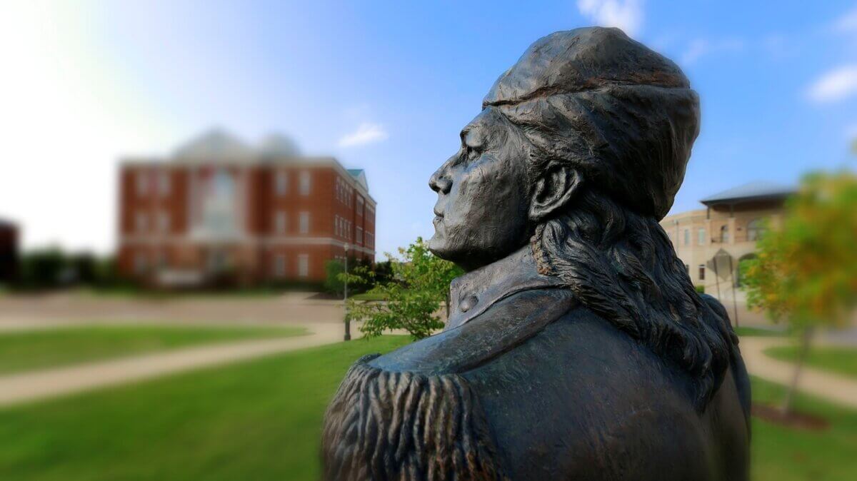 Bronze statue of a person in historical military attire, seen in profile with buildings and greenery in the background on a sunny day—a unique travel highlight during your summer getaway to Tupelo MS.