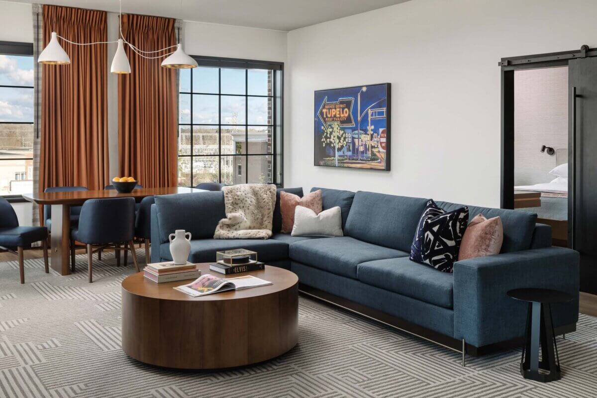 Modern living room with a blue sectional sofa, round wooden coffee table, books, and dining area with navy chairs—perfect for relaxing after a summer getaway or travel adventure in Tupelo, MS; large windows with rust-colored curtains frame the background.