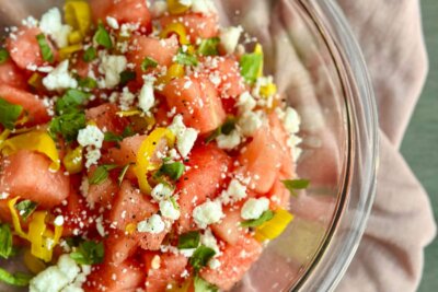 Glass bowl with diced watermelon, crumbled feta cheese, sliced yellow peppers, pepperoncini, chopped fresh herbs, and black pepper salad on a pink cloth.