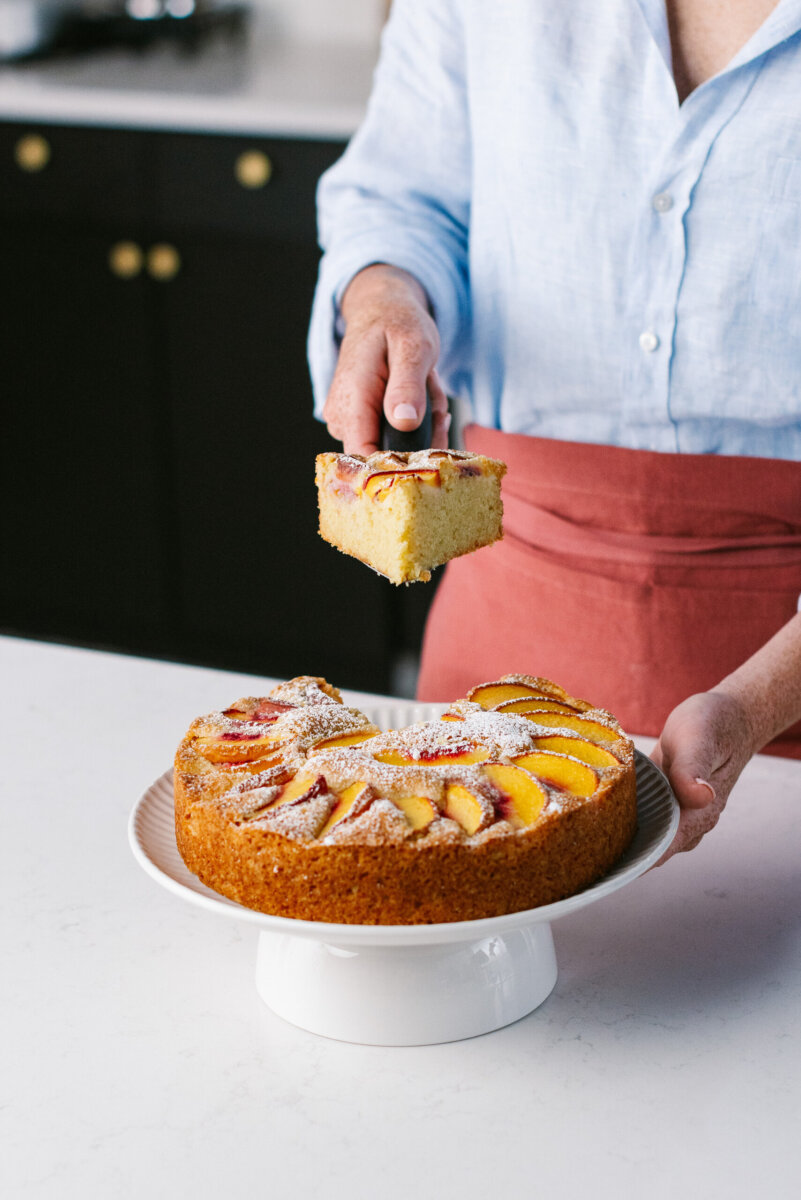 Nashville Peach Week: Limited-Time Dishes + 2 Juicy Recipes to Try at Home - 2 A person in a blue shirt and red apron lifts a slice of peach-topped olive oil cake from a whole cake on a white pedestal stand.