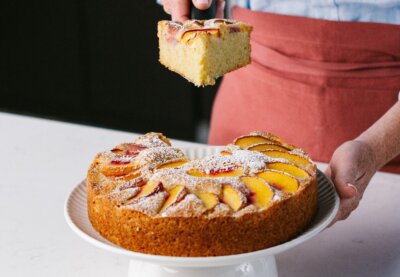 A person holds a slice of peach olive oil cake topped with powdered sugar above the remaining cake on a white cake stand.