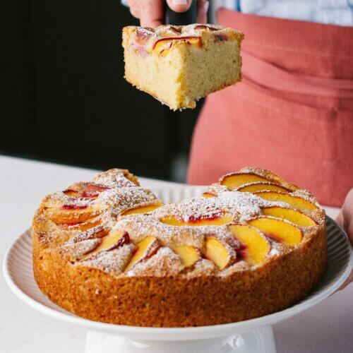 A person holds a slice of peach olive oil cake topped with powdered sugar above the remaining cake on a white cake stand.