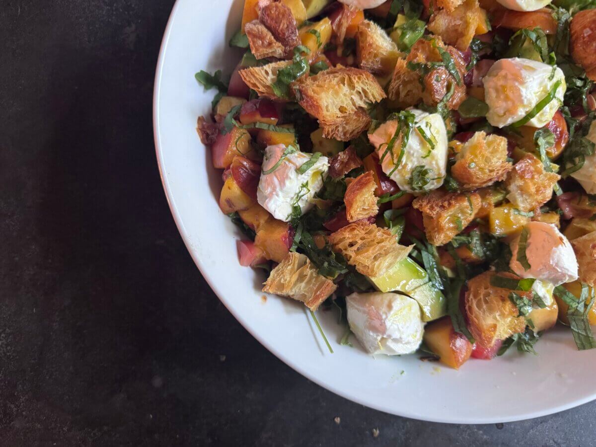 A white bowl filled with Burrata Salad featuring chunks of bread, leafy greens, diced vegetables, and dollops of soft cheese on a dark surface.