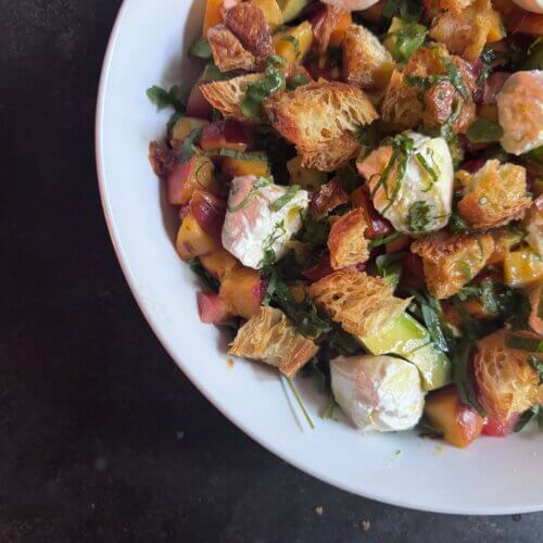 A white bowl filled with Burrata Salad featuring chunks of bread, leafy greens, diced vegetables, and dollops of soft cheese on a dark surface.