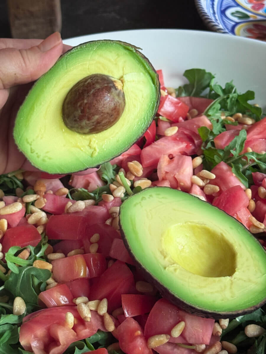 A hand holds half an avocado above a Burrata Salad with arugula, chopped tomatoes, and pine nuts in a white bowl.