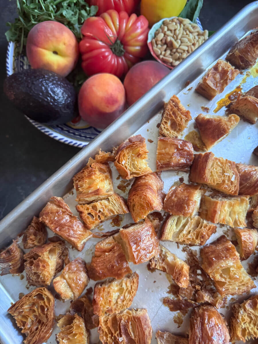 A baking tray with cubed Croissant Croutons sits next to a plate of peaches, avocado, heirloom tomato, fresh herbs, pine nuts, and a lemon—perfect for topping a Grilled Peach Salad.