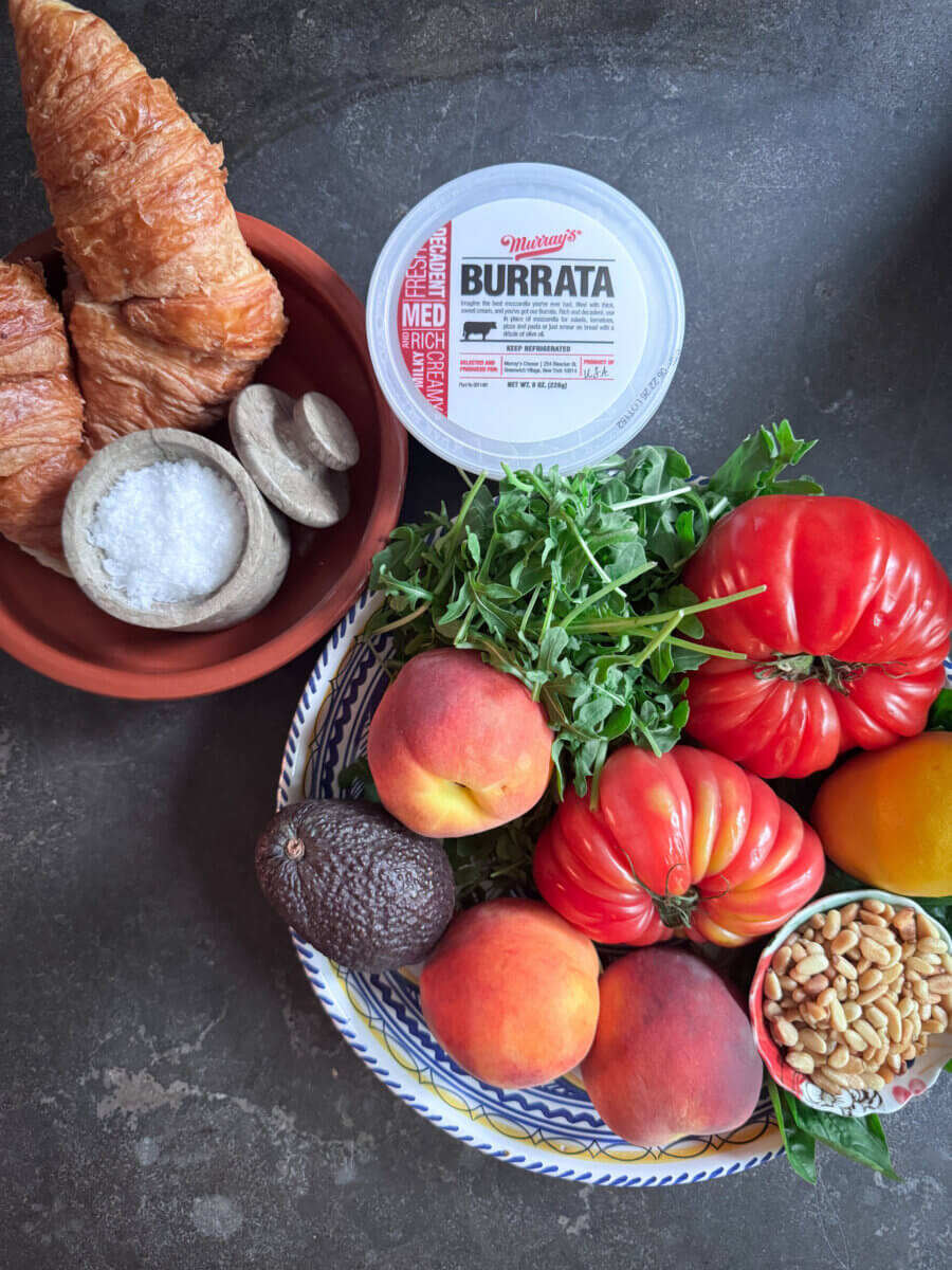 A plate with arugula, heirloom tomatoes, peaches, avocado, and pine nuts next to a container of burrata hints at a fresh Burrata Salad, served with two croissants and a small bowl of salt on a dark surface.