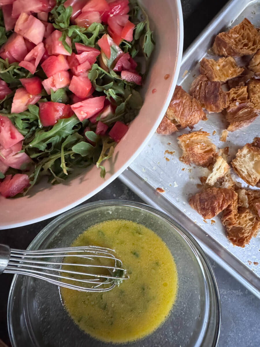 A bowl of arugula and chopped tomatoes, a tray of Croissant Croutons, and a bowl of whisked dressing with herbs sit on the kitchen counter.