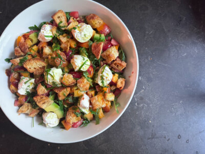 A bowl of Burrata Salad with chopped vegetables, greens, pieces of toasted bread, and dollops of soft cheese, arranged on a dark surface.