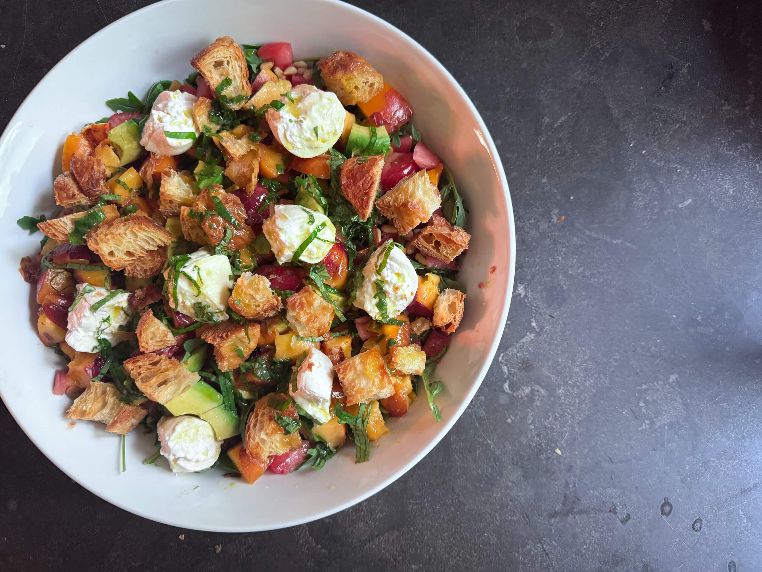 A bowl of Burrata Salad with chopped vegetables, greens, pieces of toasted bread, and dollops of soft cheese, arranged on a dark surface.