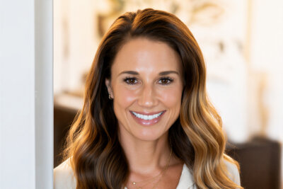 Erin Tolbert, a woman with long, wavy brown hair and a light-colored blouse, smiles at the camera in a softly lit indoor setting.