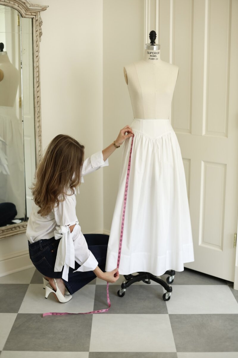 Meet Laura Finley Hasenbank, Nashville's Emerging Fashion Designer - 5 Laura Finley kneels on the floor, measuring the length of a white skirt on a dress form with a pink measuring tape in a well-lit room.