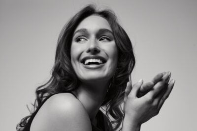 Black and white photo of Laura Finley, a young woman with wavy hair smiling and looking to the side, hands lightly clasped near her face.