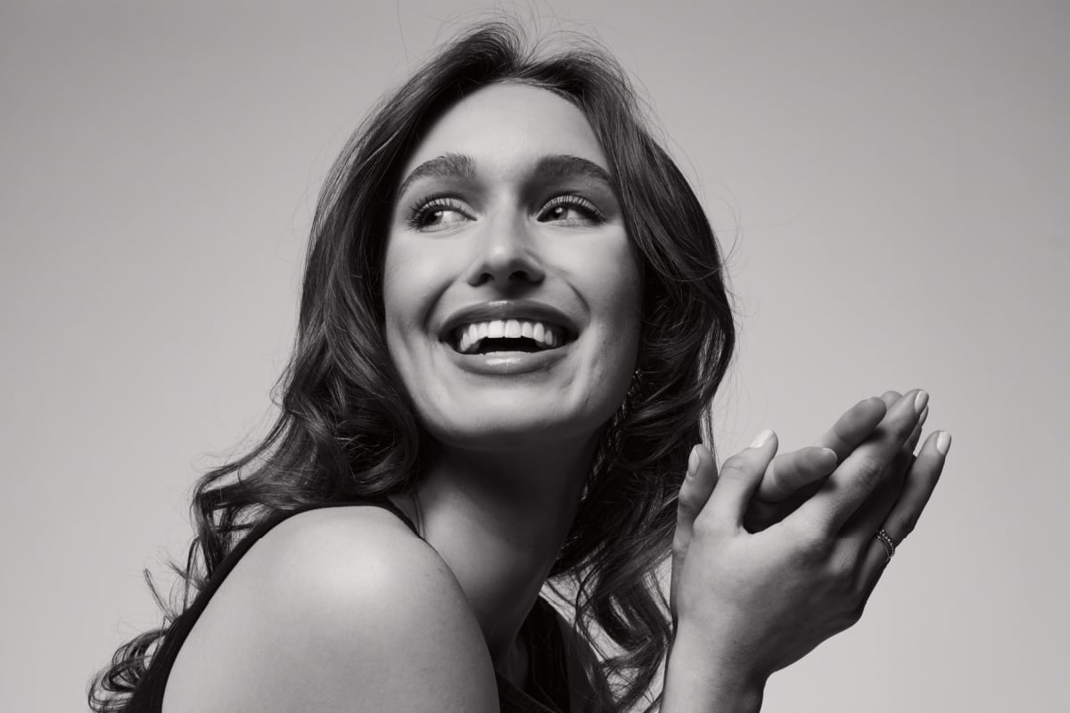 Black and white photo of Laura Finley, a young woman with wavy hair smiling and looking to the side, hands lightly clasped near her face.