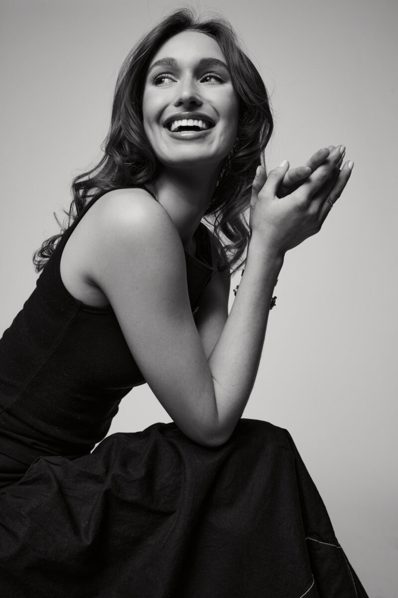 Meet Laura Finley Hasenbank, Nashville's Emerging Fashion Designer - 4 Laura Finley, with wavy hair, sits sideways, smiling and clasping her hands together, dressed in a sleeveless black top and skirt against a plain background.