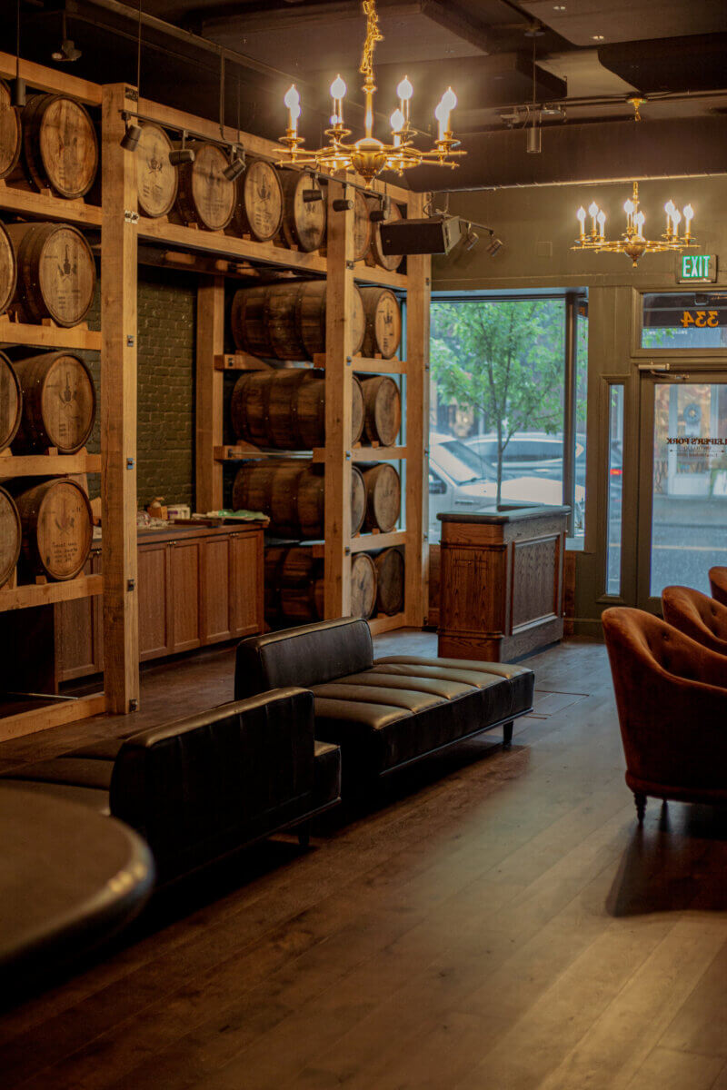 From Farm Life to Fine Spirits: Meet the Co-Owner of Leiper's Fork Distillery - 3 A bar interior inspired by Leiper's Fork Distillery, featuring wooden barrels stacked on shelves, black leather seating, chandeliers, and large windows facing the street.