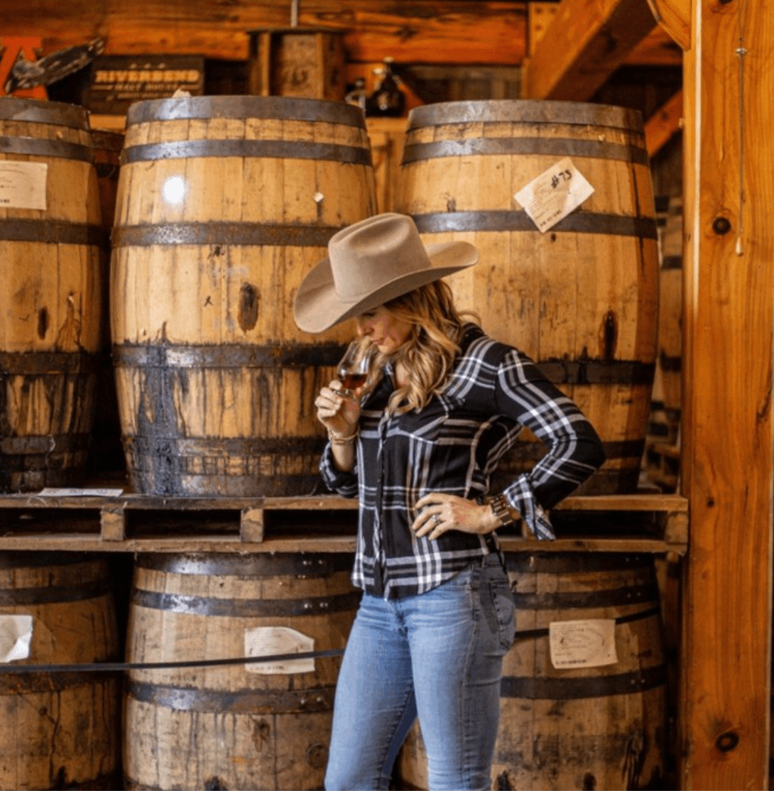 From Farm Life to Fine Spirits: Meet the Co-Owner of Leiper's Fork Distillery - 2 A person in a cowboy hat and plaid shirt stands in front of wooden barrels, smelling a drink in a glass inside the rustic Leiper's Fork Distillery.