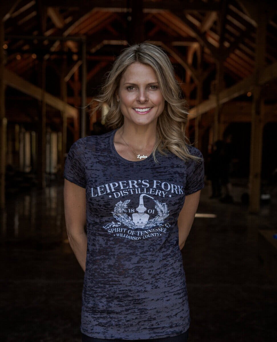 From Farm Life to Fine Spirits: Meet the Co-Owner of Leiper's Fork Distillery - 6 A woman, possibly Lynlee Kennedy, stands indoors wearing a dark Leiper's Fork Distillery t-shirt, smiling at the camera with wooden beams visible in the background.