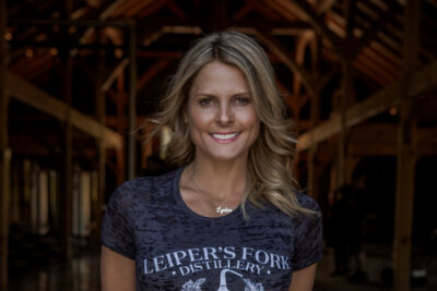 A woman with wavy blonde hair smiles at the camera, wearing a “Leiper’s Fork Distillery” T-shirt inside a wooden structure—possibly Lynlee Kennedy enjoying a moment at this iconic Tennessee distillery.
