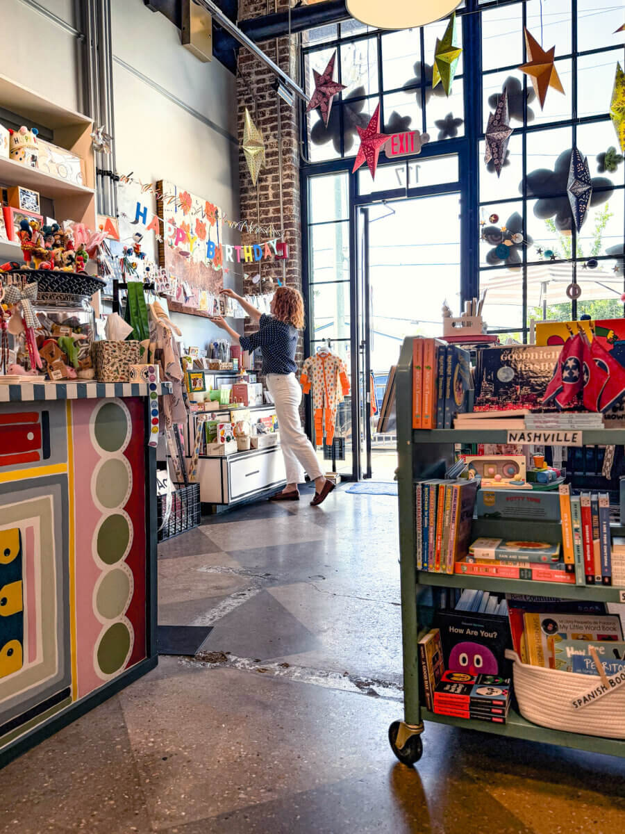 A person stands near a birthday display inside the brightly lit growUP Kids gift shop, with books and assorted items on shelves and large windows in the background.