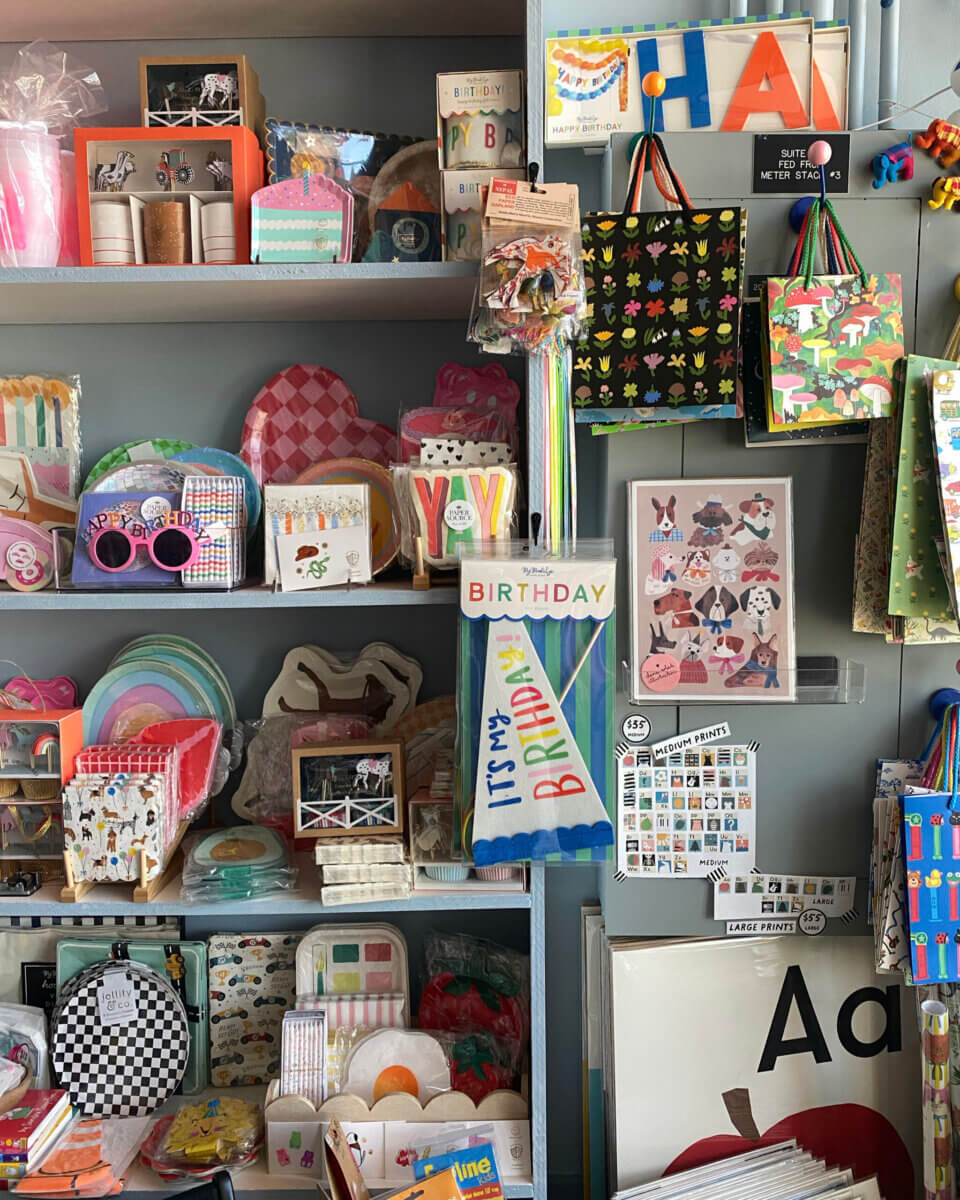 Shelves display colorful greeting cards, gift bags, party supplies, and stationery items from growUP Kids, including birthday cards, plates, napkins, and novelty sunglasses in a neatly arranged shop.