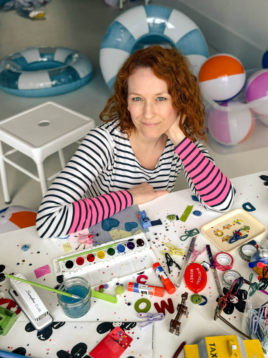 Jennifer Caywood, founder of growUP Kids, sits at a table covered with art supplies, craft materials, and toys. She wears a striped shirt and has curly hair, while inflatable pool floats and stools are visible in the background.