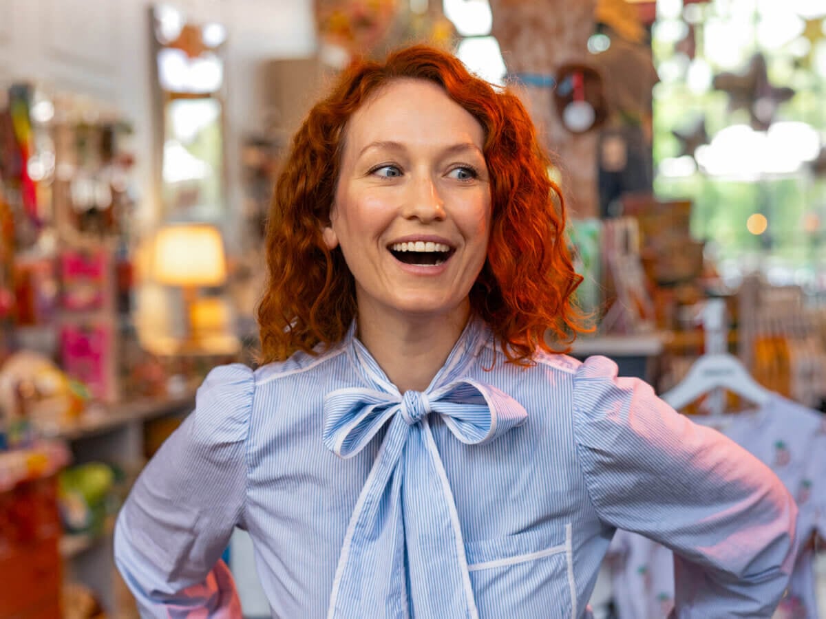 Jennifer, with her curly red hair, stands indoors in a brightly lit shop, smiling and wearing a blue blouse with a bow.