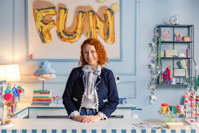 Jennifer, with her curly red hair, stands behind a counter in a colorful, decorated room featuring a “FUN” balloon sign and Grow Up FACES touches all around.