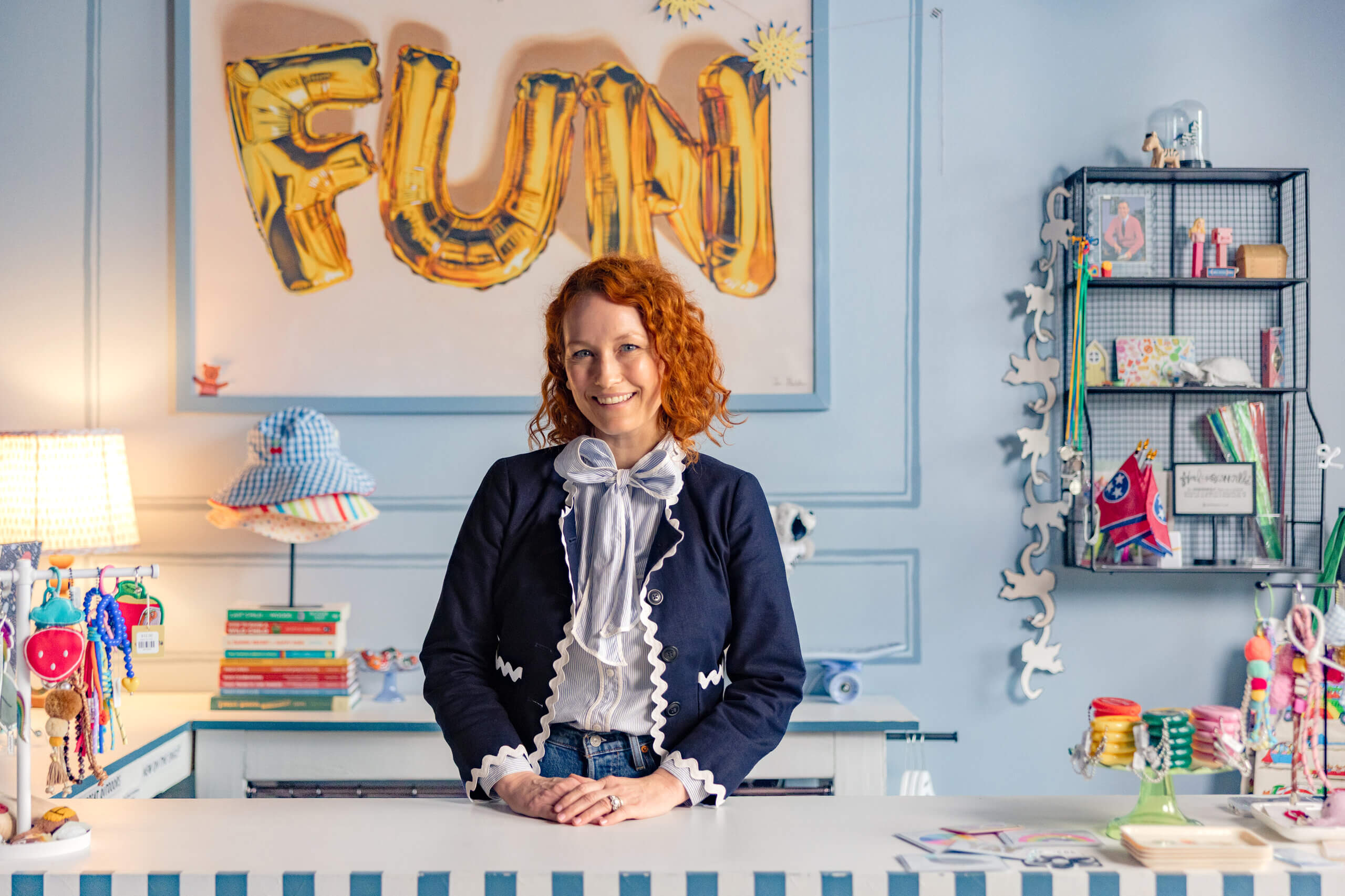 Jennifer, with her curly red hair, stands behind a counter in a colorful, decorated room featuring a “FUN” balloon sign and Grow Up FACES touches all around.