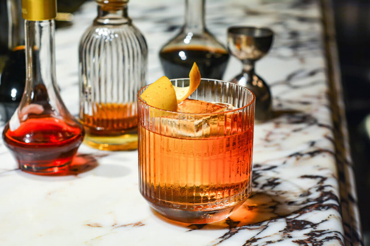 A glass of cocktail with an orange peel garnish on a marble countertop, surrounded by glass bottles and a metal jigger.