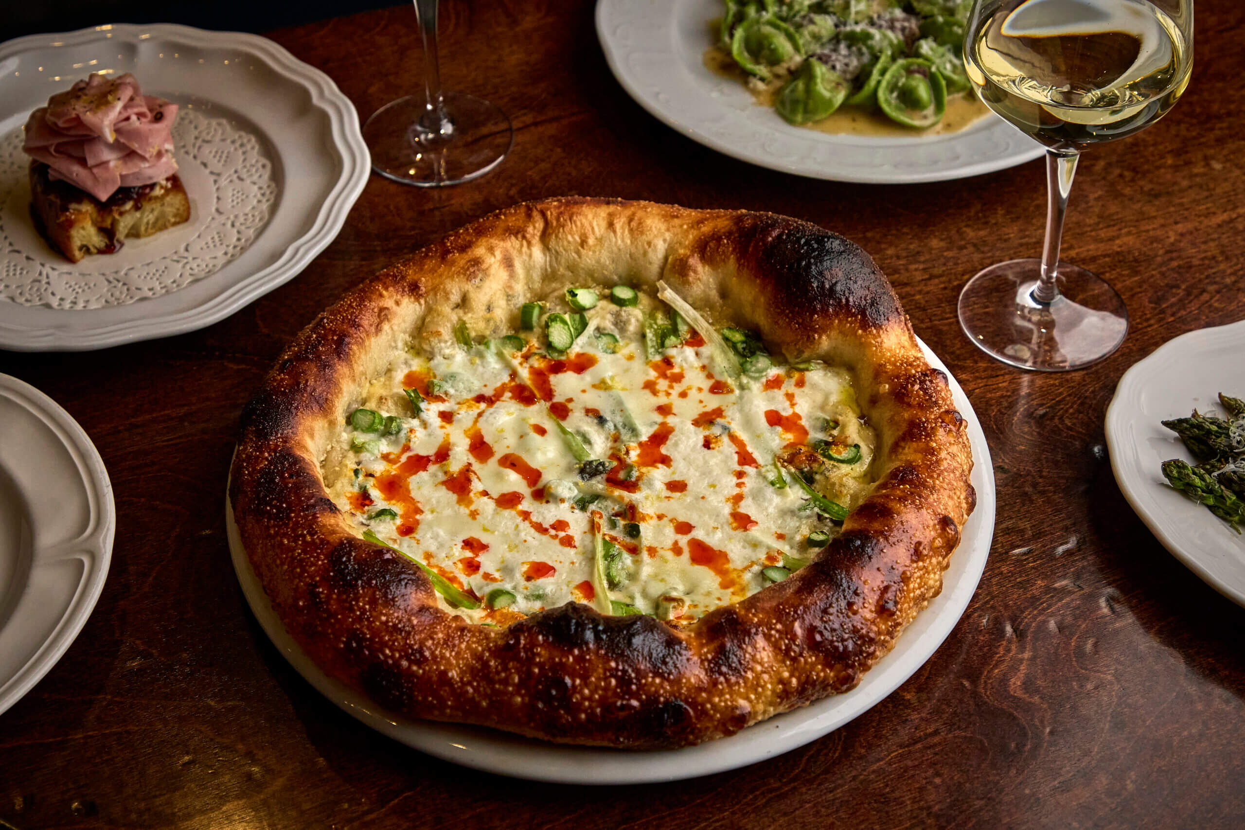 A wood-fired pizza with a bubbly, charred crust, topped with cheese, green vegetables, and red sauce—served on a white plate with wine and other dishes—highlights the vibrant flavors found at new Nashville restaurants in June 2026.