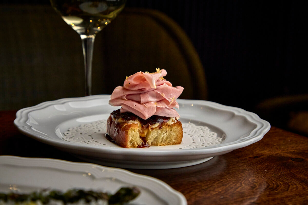 A slice of bread topped with mortadella is served on a white plate, with a wine glass in the background.