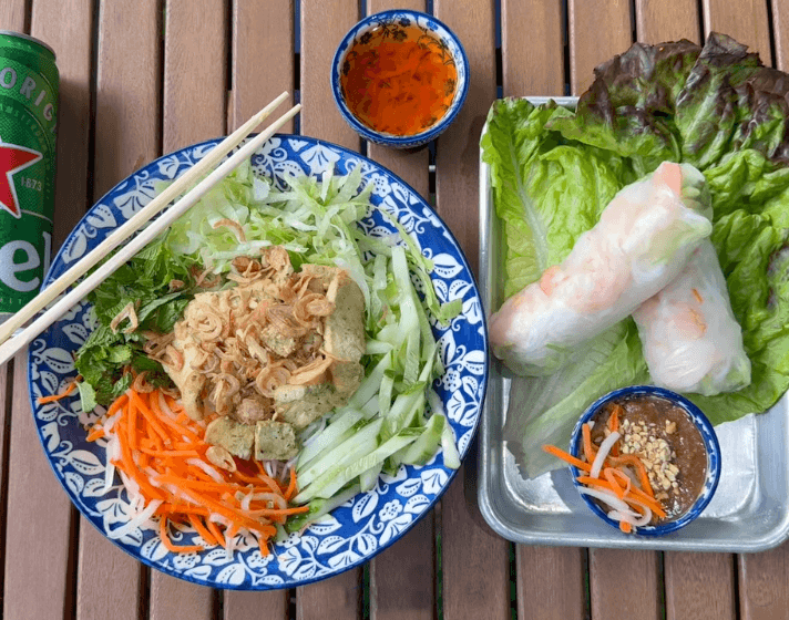 A bowl of Vietnamese noodle salad with vegetables and tofu, two fresh spring rolls with dipping sauce, and a can of Heineken on a wooden table.