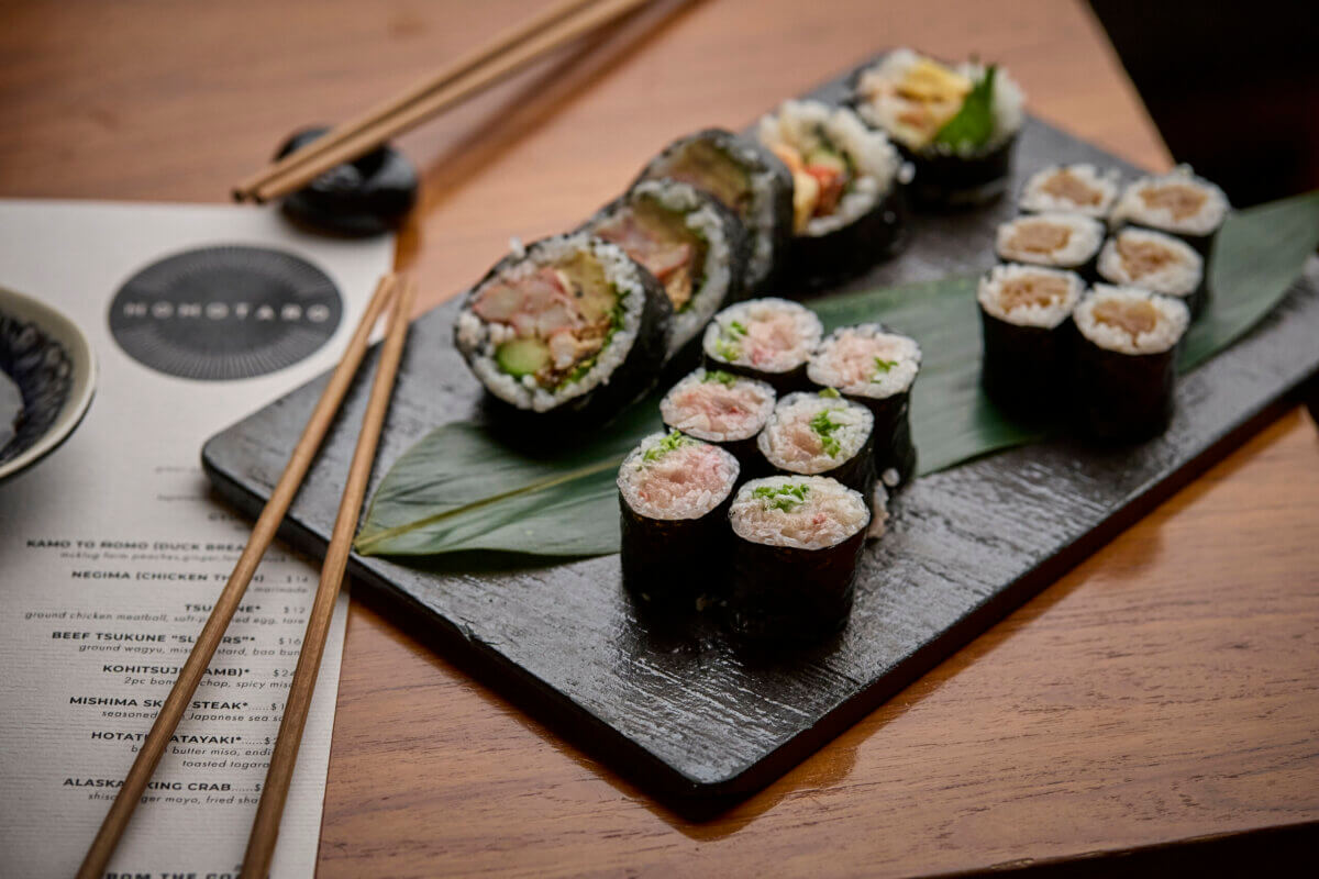 A black slate plate with assorted sushi rolls sits next to chopsticks and a printed menu on a wooden table.