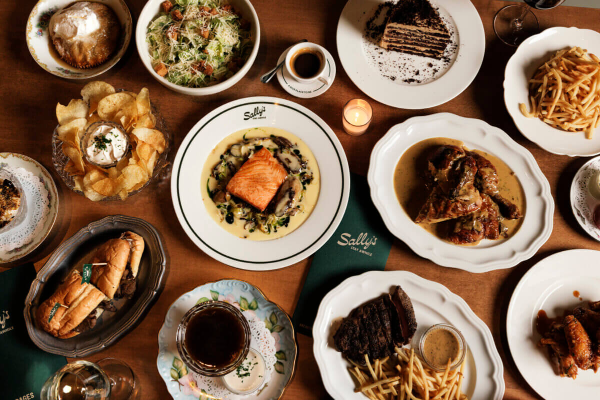 A variety of plated dishes including salmon, steak, fries, salad, dessert, and drinks are arranged on a wooden table with green napkins labeled "Sally's," one of the standout new restaurants in Nashville as of June 2026.