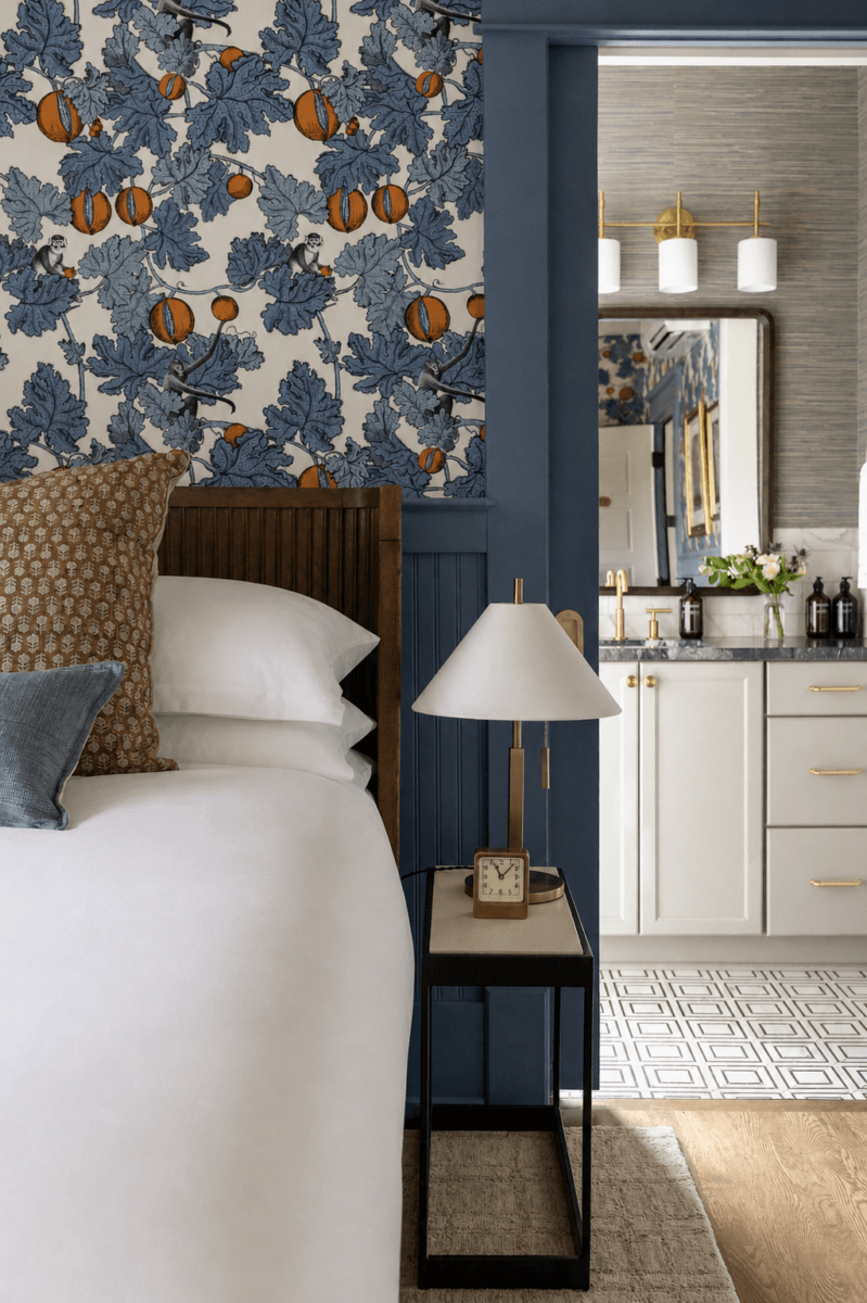 Bedroom with a wooden bed, patterned pillows, and a nightstand with a lamp and clock; blue molding and floral wallpaper; view into a modern white bathroom.