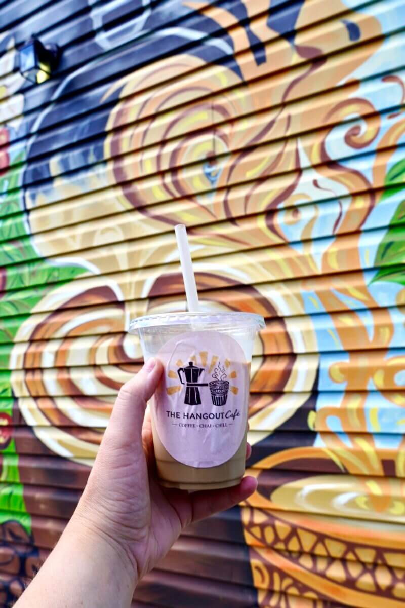 A hand holds an iced drink in a plastic cup labeled "The Hangout Cafe," one of the exciting new Nashville restaurants, in front of a colorful mural painted on a corrugated wall.