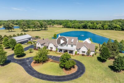 Aerial view of a large white house at 770 Ballard Rd, Piperton TN, featuring a circular driveway, lush lawns, trees, a pond with a fountain, and an outbuilding in the background.