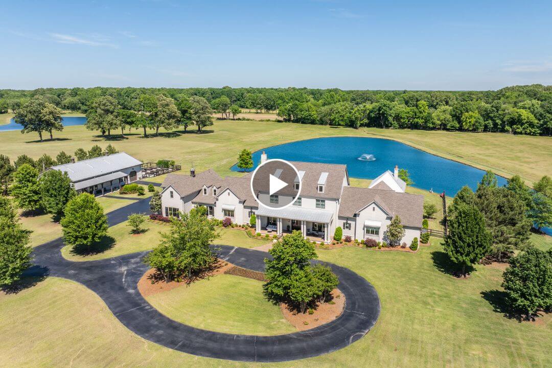 Aerial view of a large white house at 770 Ballard Rd, Piperton TN, featuring a circular driveway, lush lawns, trees, a pond with a fountain, and an outbuilding in the background.