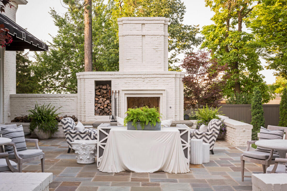 Step outside to an outdoor patio designed by Daniel Keeley, featuring a white brick fireplace, stacked firewood, a table covered with a white cloth, potted ferns, cushioned chairs, and surrounding trees that exude sophisticated style.