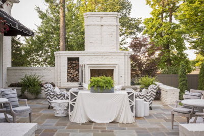 Step outside to an outdoor patio designed by Daniel Keeley, featuring a white brick fireplace, stacked firewood, a table covered with a white cloth, potted ferns, cushioned chairs, and surrounding trees that exude sophisticated style.
