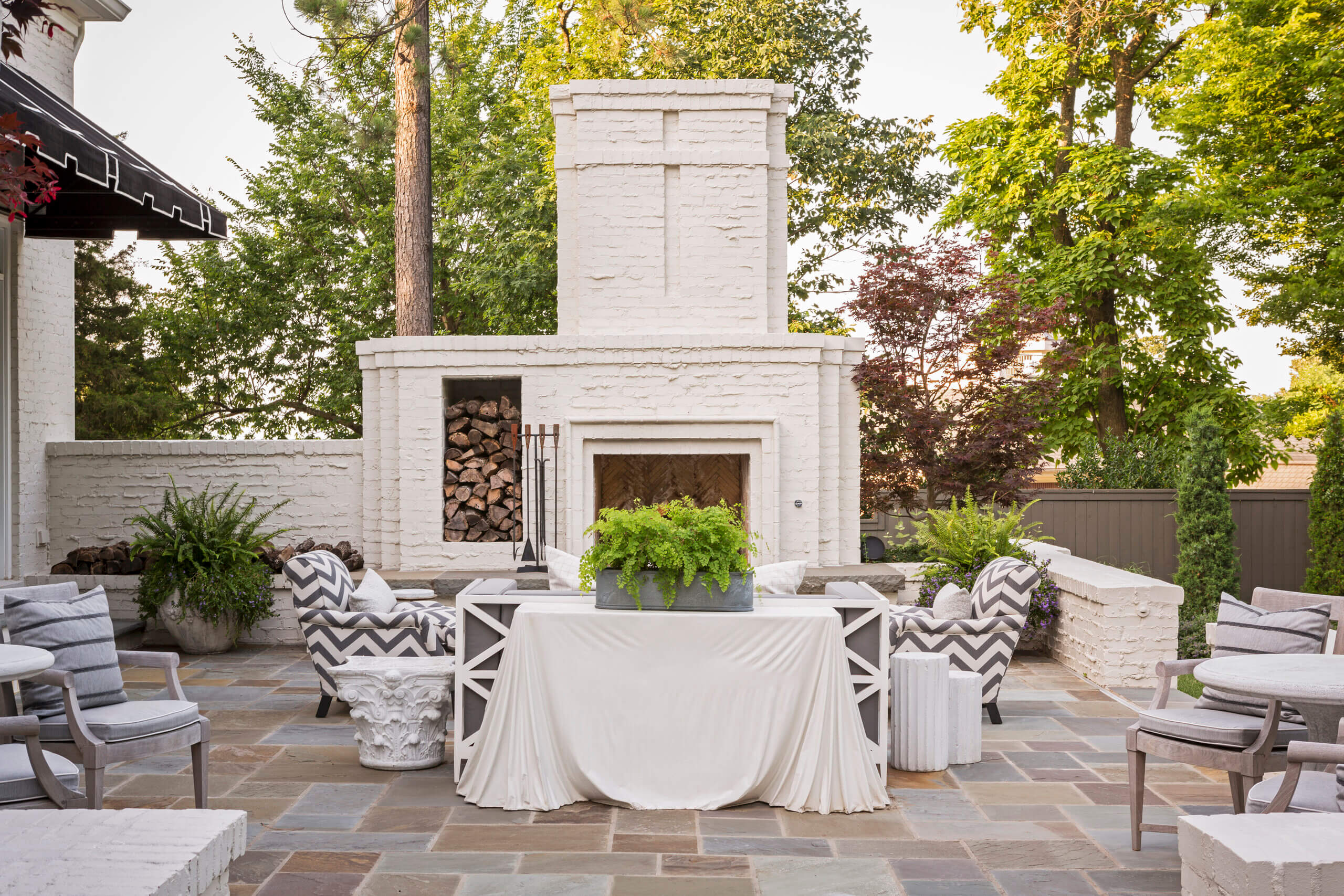 Step outside to an outdoor patio designed by Daniel Keeley, featuring a white brick fireplace, stacked firewood, a table covered with a white cloth, potted ferns, cushioned chairs, and surrounding trees that exude sophisticated style.